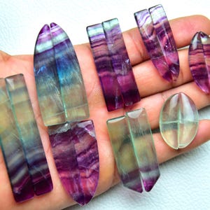 Puede incluir: Una colecci&oacute;n de piedras preciosas de fluorita pulida en varias formas, incluyendo rect&aacute;ngulos, &oacute;valos y formas puntiagudas. Las piedras muestran bandas transl&uacute;cidas de colores p&uacute;rpura, azul y verde con un acabado suave, brillante y patrones.