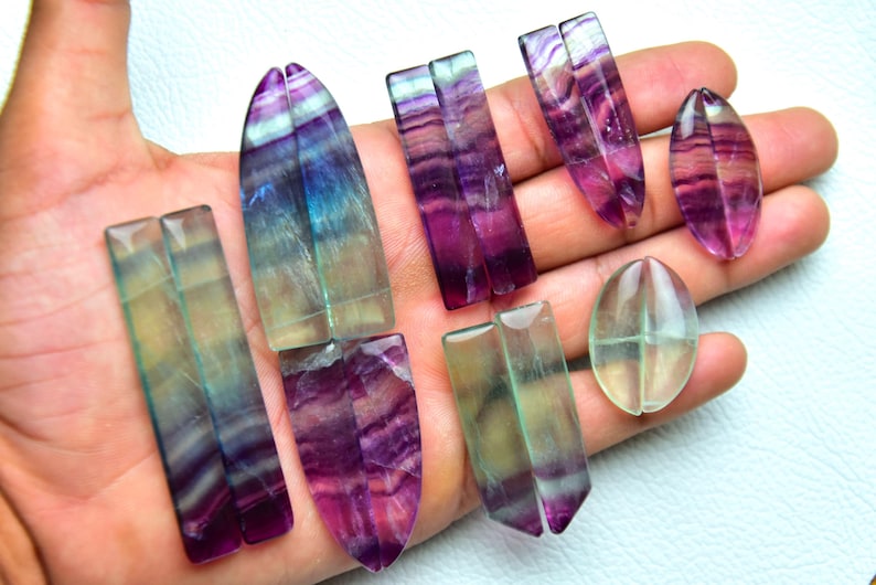 Puede incluir: Una colecci&oacute;n de piedras preciosas de fluorita pulida en varias formas, incluyendo rect&aacute;ngulos, &oacute;valos y formas puntiagudas. Las piedras muestran bandas transl&uacute;cidas de colores p&uacute;rpura, azul y verde con un acabado suave, brillante y patrones.