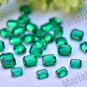 Pode incluir: Um conjunto de pedras preciosas verdes facetadas, com corte esmeralda, espalhadas sobre uma superfície branca. As pedras transparentes refletem a luz, mostrando sua cor vibrante e clareza. Ideal para joias ou artesanato.