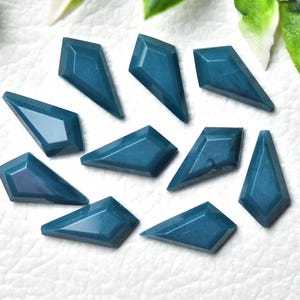 May include: A collection of ten teal-colored gemstone cabochons, each cut into a unique geometric shape with flat backs and faceted edges. The gemstones are arranged on a white surface, showcasing their polished surfaces and varying angles.
