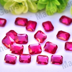 Pode incluir: Uma coleção de gemas vermelho rubi, de corte retangular e radiante. As gemas estão espalhadas sobre uma superfície branca, com algumas folhas verdes e amarelas no fundo. As gemas são facetadas, captando a luz e mostrando um aspeto brilhante.