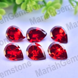 May include: Six vibrant, teardrop-shaped, faceted gemstones in a rich, deep red hue. The gems are arranged on a white surface, with green foliage in the background. The gems are likely garnets, perfect for jewelry making.