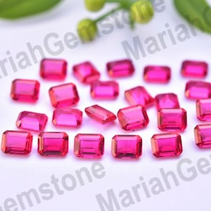 May include: A collection of radiant, rectangular-cut, hot pink gemstones. The gems are scattered on a white surface, with a few green leaves in the background. The gems are likely for jewelry making.