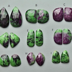 May include: A collection of polished gemstones in diverse shapes, such as teardrop, oval, and irregular forms. The stones exhibit a blend of green and purple shades, with some showing black speckling. The gemstones are displayed on a light grey surface.