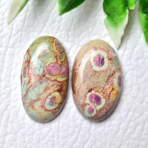 May include: Two oval-shaped gemstones with unique patterns. One features a mix of brown, green, and pink hues, while the other has a similar color palette with a lighter base. The stones are set against a white background.