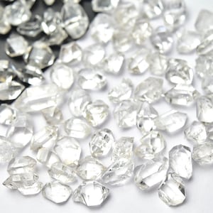 Może przedstawiać: Kolekcja przezroczystych, nieoszlifowanych diamentów rozrzuconych na białej powierzchni. Diamenty różnią się rozmiarem i kształtem, z wieloma fasetami odbijającymi światło. Niektóre są trzymane w czarnej tacy, inne są luźne.