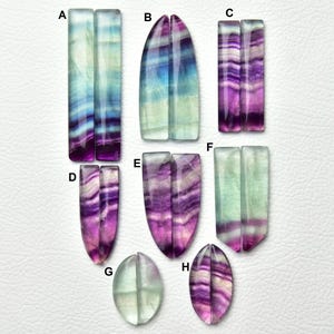 Puede incluir: Una colecci&oacute;n de pares de piedras preciosas de fluorita pulida etiquetadas de la A a la H, con patrones de bandas en tonos morado, azul, verde y blanco. Las piedras tienen formas rectangulares, ovaladas alargadas y de l&aacute;grima con superficies lisas.