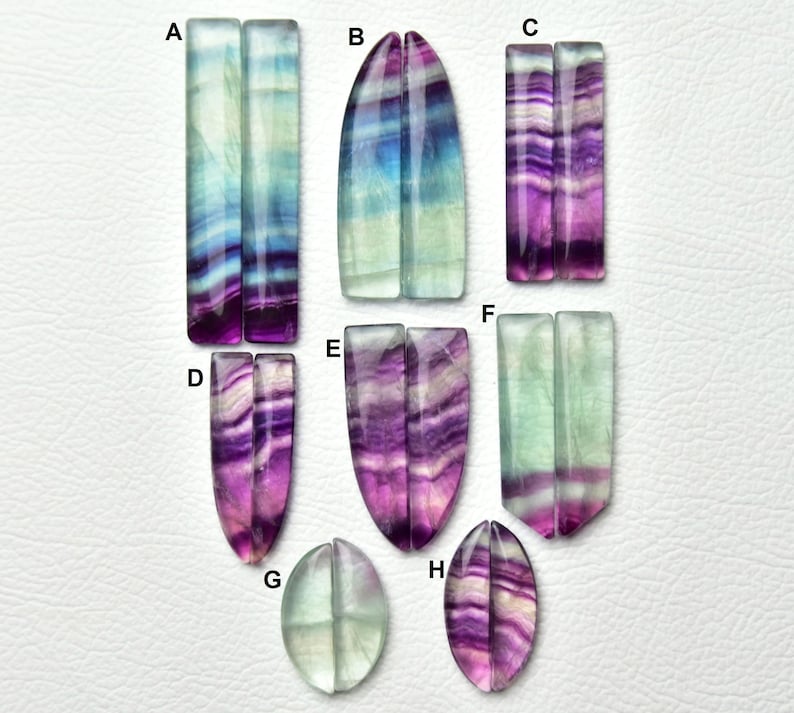 Puede incluir: Una colecci&oacute;n de pares de piedras preciosas de fluorita pulida etiquetadas de la A a la H, con patrones de bandas en tonos morado, azul, verde y blanco. Las piedras tienen formas rectangulares, ovaladas alargadas y de l&aacute;grima con superficies lisas.