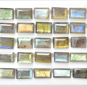 May include: A collection of twenty-five rectangular labradorite gemstones arranged in a five-by-five grid. Each stone features a faceted surface with iridescent flashes of blue, gold, green, and grey tones, displaying natural variations in color.