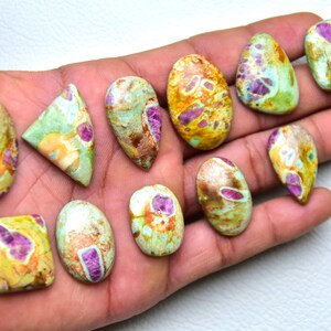 May include: A collection of eleven polished cabochons featuring a mix of green, yellow, and brown matrix stone with embedded magenta-colored inclusions. The stones are shaped in various ovals, triangles, and rectangles with smooth surfaces.