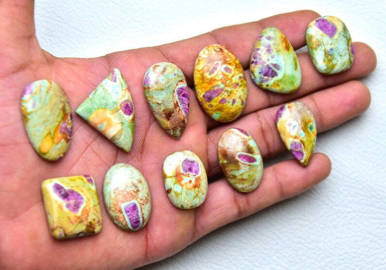 May include: A collection of eleven polished cabochons featuring a mix of green, yellow, and brown matrix stone with embedded magenta-colored inclusions. The stones are shaped in various ovals, triangles, and rectangles with smooth surfaces.