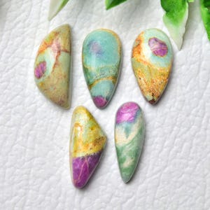 May include: Five polished gemstones in various shapes. The stones display a mix of colours, including green, yellow, purple, and teal. Each stone has a unique pattern and texture, with a polished, smooth surface. The gemstones are arranged on a white surface.