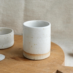 May include: A cylindrical ceramic tumbler with a speckled white glaze and a raw, unglazed tan base. Beside it sits a smaller, matching ceramic bowl. Both items feature a rustic, hand-thrown appearance with visible clay texture and earthy tones.