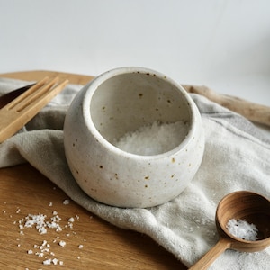May include: A small, speckled ceramic salt bowl filled with salt. A wooden spoon with salt is next to it on a wooden board. A linen cloth and wooden fork are also visible, creating a rustic kitchen aesthetic.