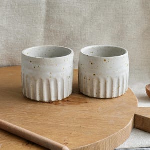 May include: Two cylindrical ceramic cups with a speckled off-white glaze and a vertical ribbed texture around the lower half. The cups have a smooth, rounded rim and a consistent, sturdy shape, showcasing a natural, earthy aesthetic in their design.