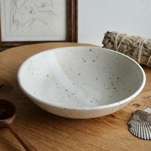 May include: A speckled white ceramic bowl with a rounded shape. The bowl has small brown spots and sits on a wooden surface. A wooden spoon, a seashell, and a bundle of sage are also visible.