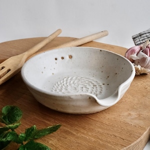Könnte beinhalten: Eine gesprenkelte, weiße Knoblauchreibe aus Keramik mit einer strukturierten Reibefläche. Die Schüssel hat einen kleinen Ausguss und drei Löcher. Eine Holzgabel und eine Knoblauchzehe befinden sich im Hintergrund. Die Schüssel steht auf einem Holzschneidebrett.
