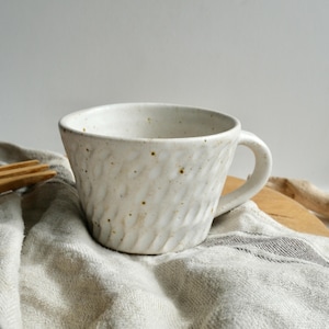 May include: A textured, off-white ceramic mug with a handle. The mug has a slightly irregular shape and a speckled surface. The mug is sitting on a light-coloured, striped cloth.