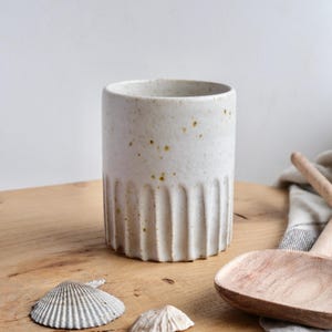 Könnte beinhalten: Ein zylindrischer, cremefarbener Keramikbecher mit vertikalen Rippendetails im unteren Bereich. Der Becher hat kleine braune Sprenkel und steht auf einer Holzoberfläche mit Muscheln und einem Holzlöffel. Für Küche oder Esszimmer.