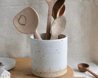 May include: A white ceramic utensil holder with brown speckles containing several wooden spoons of varying shapes and sizes. The holder is cylindrical with a smooth finish and a natural clay base. Additional wooden spoons and small ceramic items are nearby.