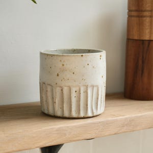 May include: A cream-coloured ceramic cup with vertical ribbed detailing on the lower half. The cup sits on a wooden shelf, with a wooden object in the background. The cup is a neutral colour and could be used for a variety of purposes.