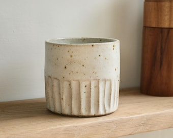 May include: A speckled, off-white ceramic cup with vertical ribbed detailing on the lower half. The cup sits on a wooden shelf, with a wooden object in the background. The cup is a neutral color and could be used for a variety of purposes.