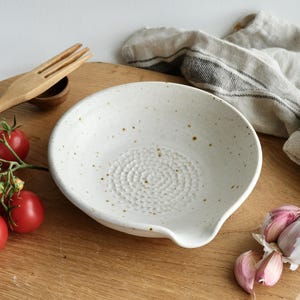 May include: A speckled white ceramic garlic grater bowl with a textured grating surface and a pouring spout. The bowl is on a wooden board with tomatoes, garlic, and a wooden fork. The bowl is for kitchen use.
