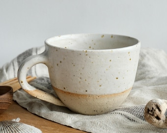 May include: A speckled white ceramic mug with a tan base and handle. The mug is sitting on a light gray and white striped cloth, with a wooden cutting board and seashell in the background. The mug has a rustic, handmade appearance.