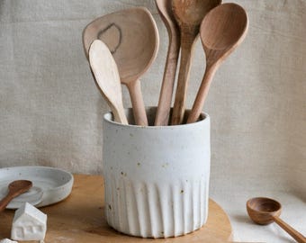 May include: A white ceramic utensil holder with vertical fluted sides and brown speckles, containing five wooden spoons of varying shapes and sizes. A small white ceramic house figurine and a wooden spoon rest are placed nearby.