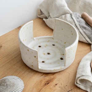 May include: A speckled, off-white ceramic soap dish with a circular design and cut-out sides. The dish has drainage holes and sits on a wooden surface. A seashell and linen towels are also visible.