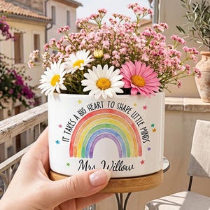 May include: A white ceramic planter with a wooden base, featuring a colorful watercolor rainbow graphic and the text "It takes a big heart to shape little minds" and "Mrs. Willow" in black script, filled with pink baby's breath and white daisies.