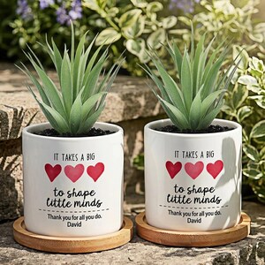 May include: Two white ceramic cylindrical planters, each holding a green succulent plant and resting on a circular bamboo saucer. The planters feature the printed text: It takes a big heart to shape little minds. Thank you for all you do. David.