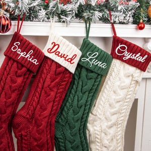 May include: Four cable-knit Christmas stockings featuring folded cuffs. The first is red with white embroidery reading Sophia, the second is red with white cuffs and red embroidery reading David, the third is green with white embroidery reading Luna, and the fourth is white with red cuffs and white embroidery reading Crystal.