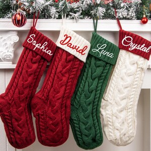 May include: Four cable-knit Christmas stockings hanging in a row. Two are red with white embroidered names Sophia and David, one is green with the white embroidered name Luna, and one is white with the red embroidered name Crystal.