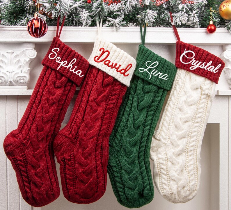 May include: Four cable-knit Christmas stockings hanging in a row. Two are red with white embroidered names Sophia and David, one is green with the white embroidered name Luna, and one is white with the red embroidered name Crystal.