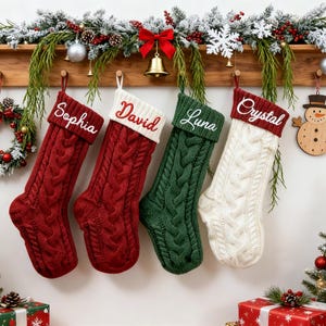May include: Four cable-knit Christmas stockings hanging from a wooden rack. Two are deep red with white cuffs, one is dark green with a white cuff, and one is white with a red cuff. Each cuff features embroidered names: Sophia, David, Luna, and Crystal.