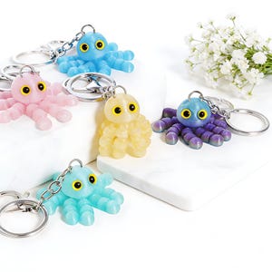 May include: A collection of colorful octopus keychains in shades of blue, pink, yellow, and purple. Each keychain features a detailed octopus design with large, yellow eyes and a silver keyring. The keychains are displayed on a white surface.