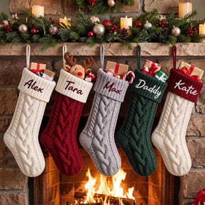 May include: Five cable-knit Christmas stockings hanging in a row. The stockings are cream, red, gray, dark green, and cream, each featuring a personalized name embroidered on the cuff: Alex, Tara, Max, Daddy, and Katie, with small gifts inside.