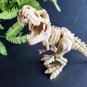 Może przedstawiać: Beżowa figurka szkieletu Tyrannosaurus Rex. Model dinozaura ma szczegółową strukturę szkieletu, w tym żebra, kręgosłup i czaszkę z widocznymi zębami. Figurka jest ustawiona w pozycji stojącej.