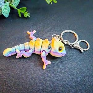 Pode incluir: Um chaveiro de lagarto articulado colorido em tons de amarelo, roxo e azul. O lagarto tem um corpo segmentado e um chaveiro e fecho prateados. O chaveiro foi projetado para se assemelhar a um camaleão ou um gecko.