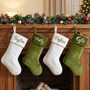 May include: Four Christmas stockings hanging in a row, alternating between white and olive green velvet fabric. Each stocking features a contrasting cuff with a personalized name embroidered in script: Taylor, Polly, Dylan, and Kyle.