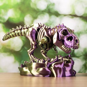 Puede incluir: Figura de esqueleto de Tyrannosaurus Rex metálico púrpura y dorado. El dinosaurio está posado sobre una base rocosa, con las mandíbulas abiertas. La figura es un artículo decorativo, probablemente para exhibición.