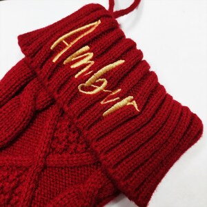 May include: A deep red knitted Christmas stocking featuring a ribbed cuff with the word Amour embroidered in gold thread. The main body of the stocking displays a textured cable knit pattern, and a matching red yarn loop is attached at the top.