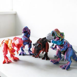 Peut inclure: Plusieurs dinosaures jouets colorés en rouge, orange, bleu et noir. Les dinosaures en plastique sont présentés avec la gueule ouverte, montrant leurs dents. Les jouets sont sur une surface blanche.
