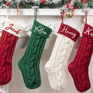 May include: Four cable-knit Christmas stockings hanging in a row. The first is red with a white cuff embroidered with the name Jacob. The second is green with a green cuff embroidered with Alex. The third is white with a red cuff embroidered with Henry. The fourth is red with a red cuff embroidered with Katie.