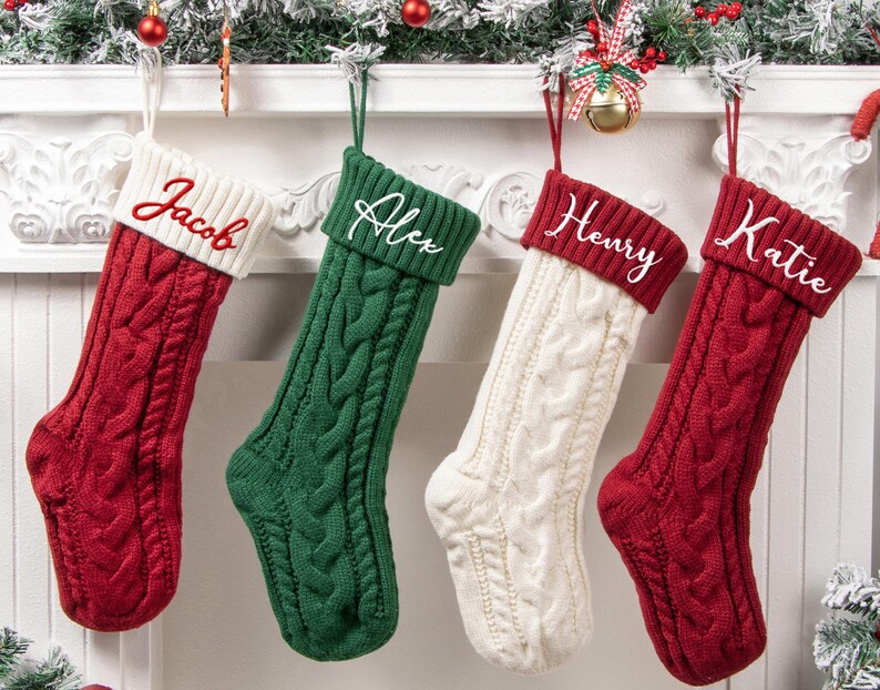 May include: Four cable-knit Christmas stockings hanging in a row. The first is red with a white cuff embroidered with the name Jacob. The second is green with a green cuff embroidered with Alex. The third is white with a red cuff embroidered with Henry. The fourth is red with a red cuff embroidered with Katie.