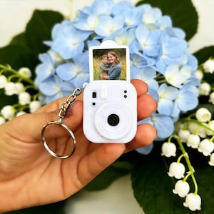 May include: A white camera-shaped keyring with a small photo of a mother and child. The keyring has a silver chain and ring. The background features blue hydrangea flowers and white lily of the valley.
