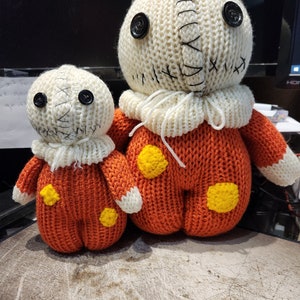 May include: Two knitted plush toys in the shape of a stitched-up doll. The larger doll is orange with yellow accents and the smaller doll is the same but smaller. Both dolls have white knitted faces with black button eyes.