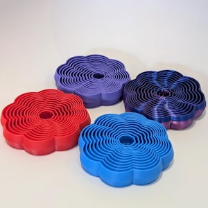 Puede incluir: Cuatro cortadores de galletas con forma de flor, uno rojo, uno azul, uno morado y uno azul oscuro. Los cortadores están superpuestos para crear un efecto 3D.