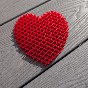 May include: A red heart-shaped object made of a grid pattern. The object is made of plastic and is sitting on a gray wooden surface.